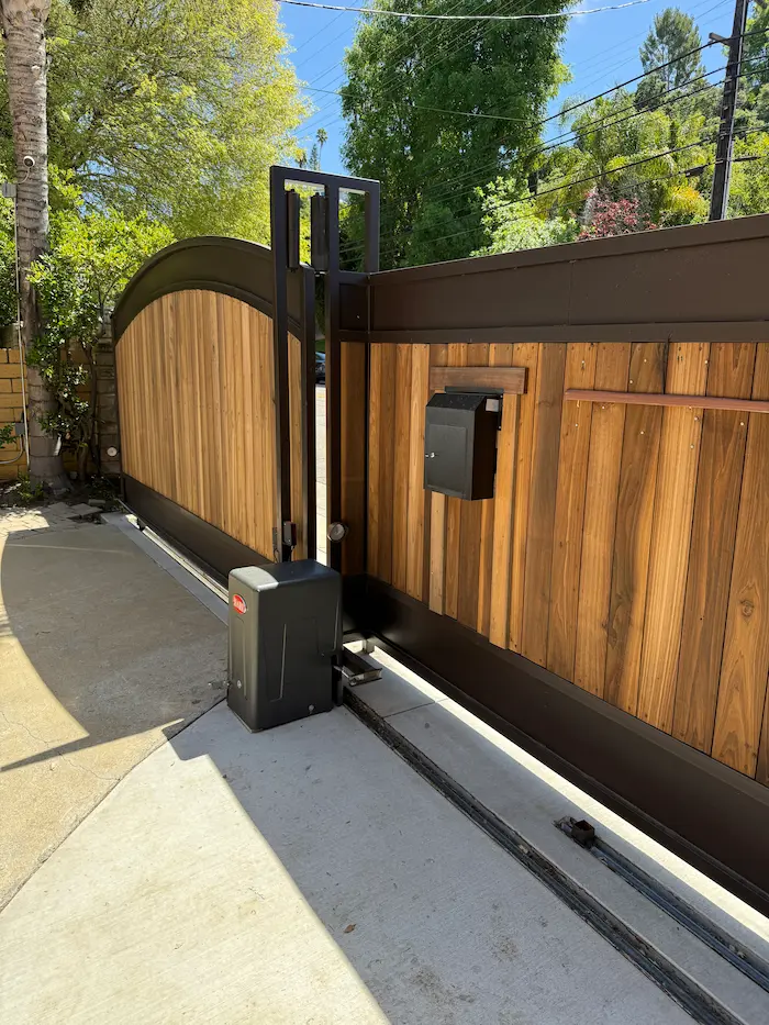 Modern sliding driveway gate with vertical wood panels and a dark metal frame. Features a curved top, built-in black mailbox, and an automatic gate motor system.