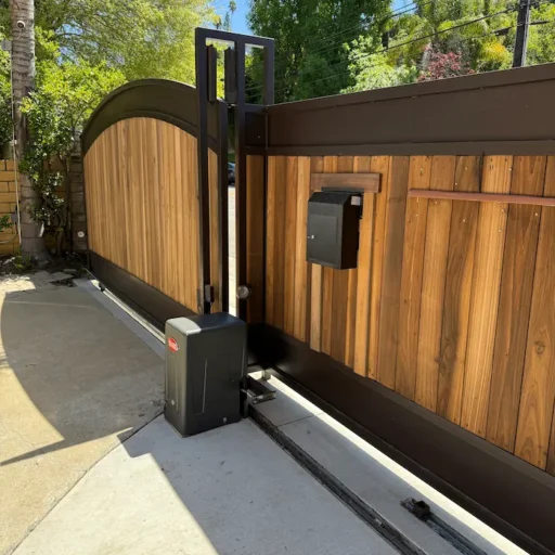 Modern sliding driveway gate with vertical wood panels and a dark metal frame. Features a curved top, built-in black mailbox, and an automatic gate motor system.