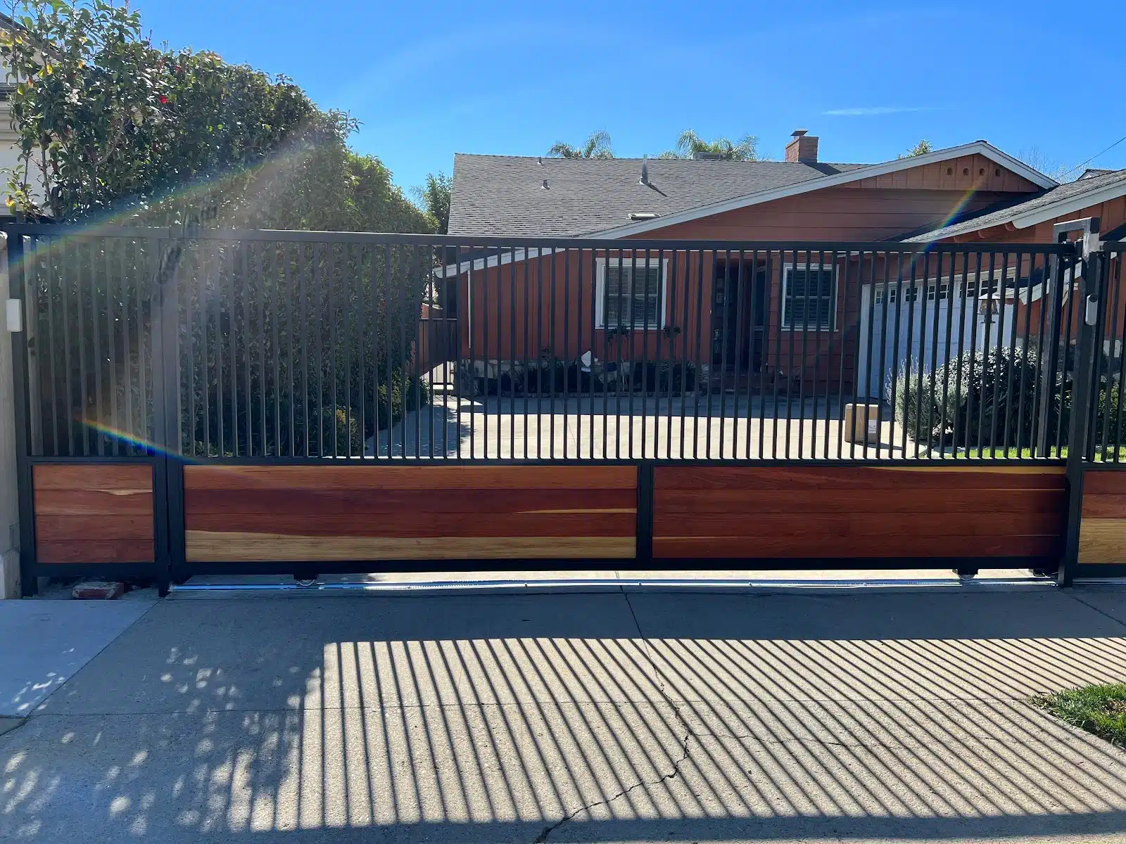 Black sliding driveway gate with vertical steel bars on top and a horizontal wood panel base, mounted on a rolling track